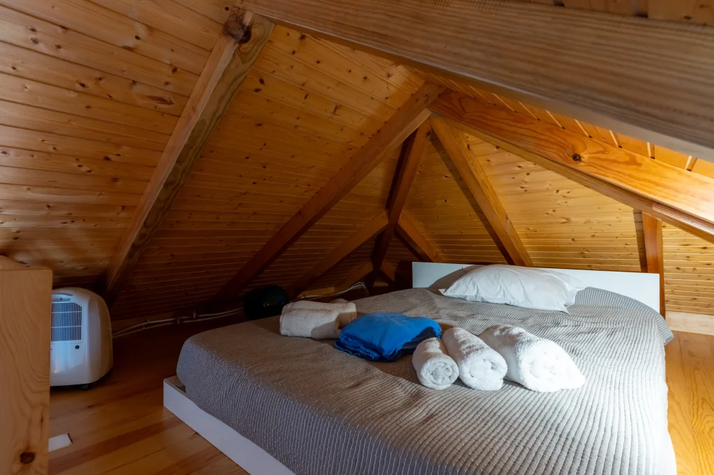 O quarto no sótão da Villa Escaleiras, sob as asnas de madeira à vista, com a cama feita e toalhas enroladas