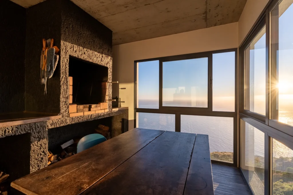 Villa Espigão's outdoor dining nook, a stone barbecue hearth beside floor-to-ceiling windows over the Atlantic