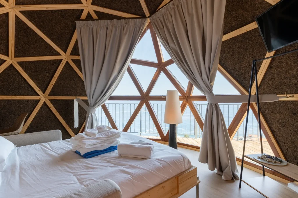 Villa Domaterra II's bed with folded towels, looking out through the dome's triangular windows to the deck and the sea, a TV on the cork wall